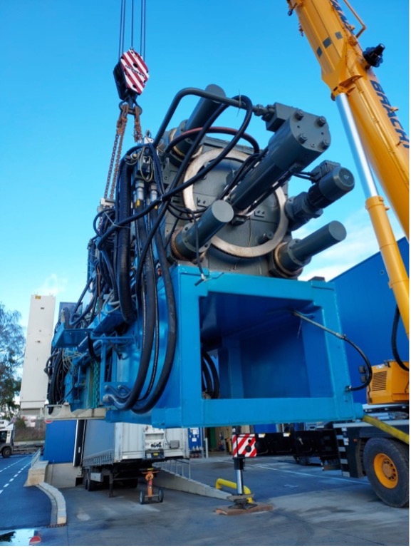 Ein Schwerlastkran hebt eine große blaue Industriemaschine mit zahlreichen Hydraulikleitungen und Zylindern in die Luft.